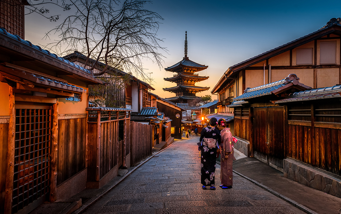 a stunning scene of Kyoto at dusk. the quiet street is lined with traditional buildings while two geisha women talk. 