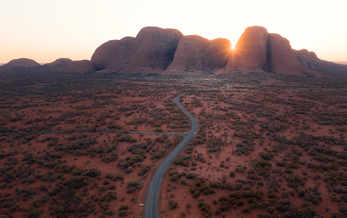 a winding road leads to the stunning red domes of Kata Tjuta. The sun peaking through the middle.