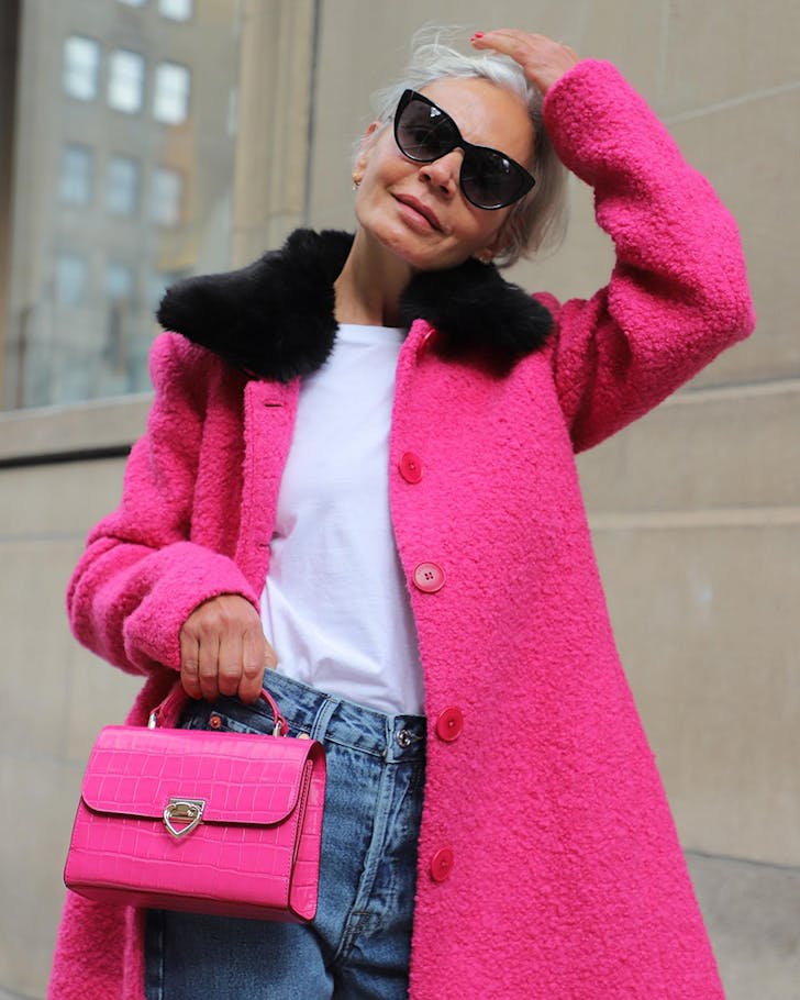 A person stands on the sidewalk dressed in a vibrant pink coat holding a vibrant pink Kate Space handbag. 