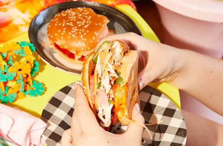 Hands wrapped around a home-made crispy fried chicken burger with a black and white paper plate underneath. 