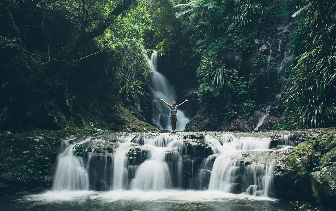 man with his hands in the air, standing on the edge of a small waterfall amongst the trees