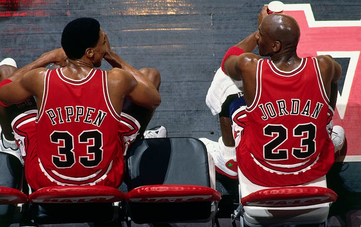 Chicago Bulls teammates Scottie Pippen and Michael Jordan sit court-side, at half-time during a game. They have their backs to the camera with their jerseys on proud display, with their heads close as they talk game play