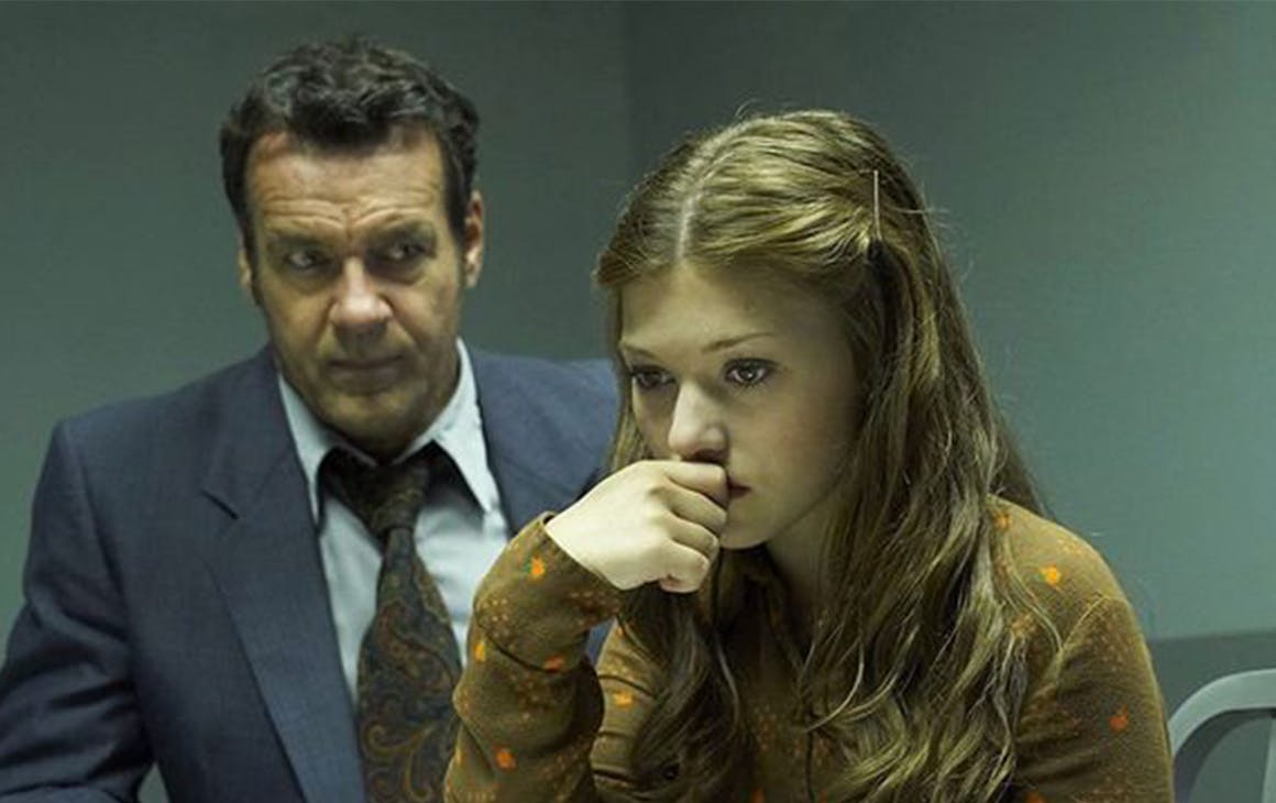 a young woman rests her head on her hand while a man in a suit sits behind her.