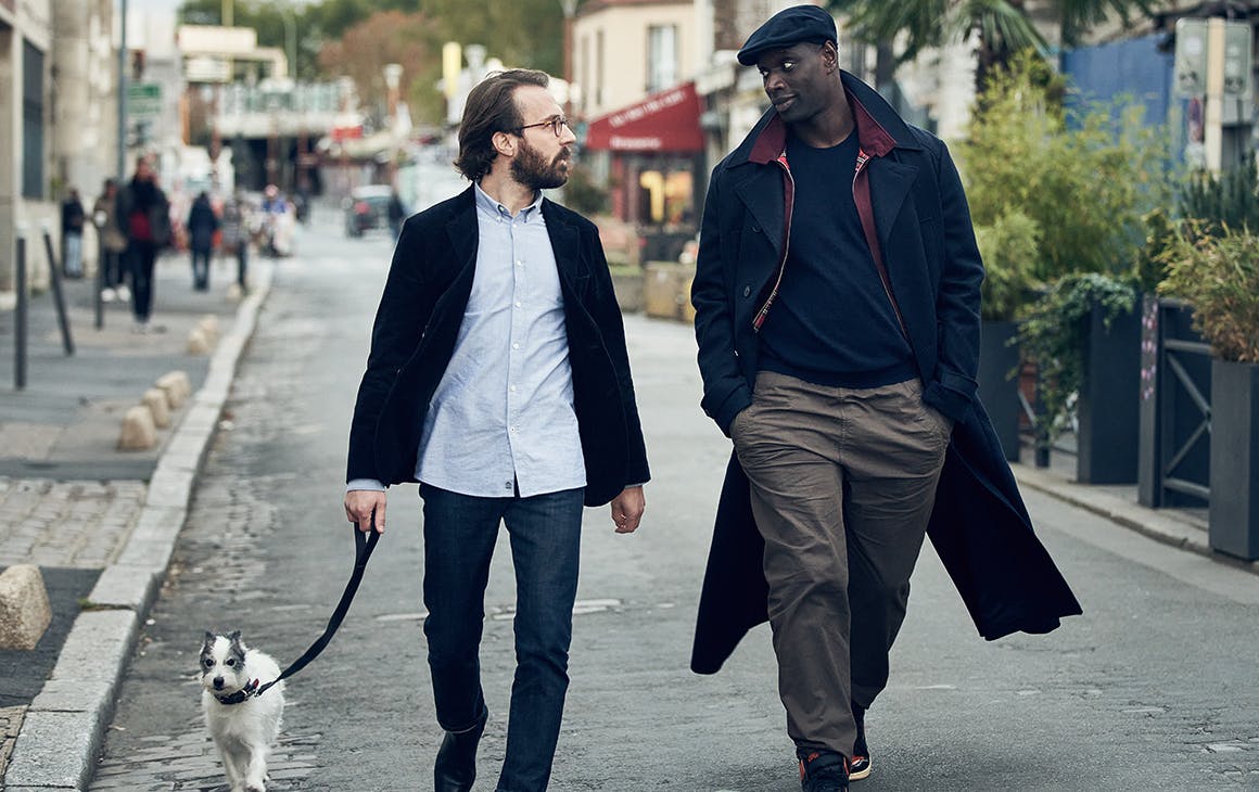 Two men, one holding a dog's leash, walk down a village street.