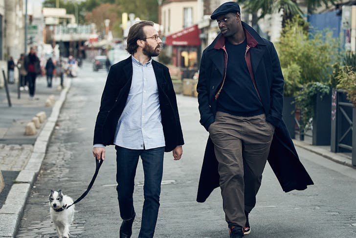 Two men, one holding a dog's leash, walk down a village street.