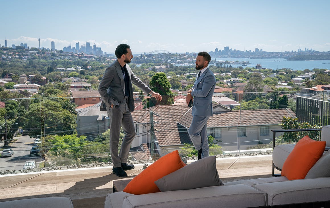 Two men in suits stand on a balcony overlooking the Sydney harbour. 