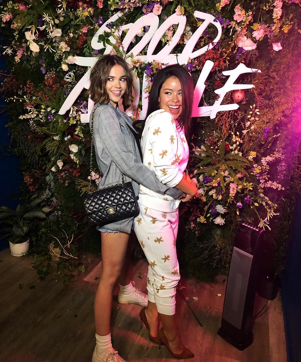 Maia Mitchell and Cierra Ramirez embrace in front of flower wall with the words Good Trouble on it.