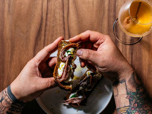 two tattooed hands holding bread with wine glass nearby