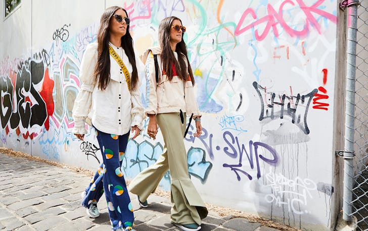 two young women with long hair walk down an alley way with graffiti 