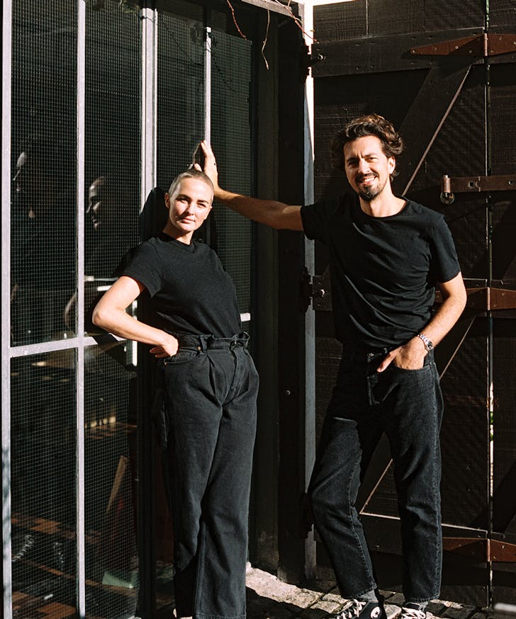 a young man and woman, dressed in black, lean against a wire fence.