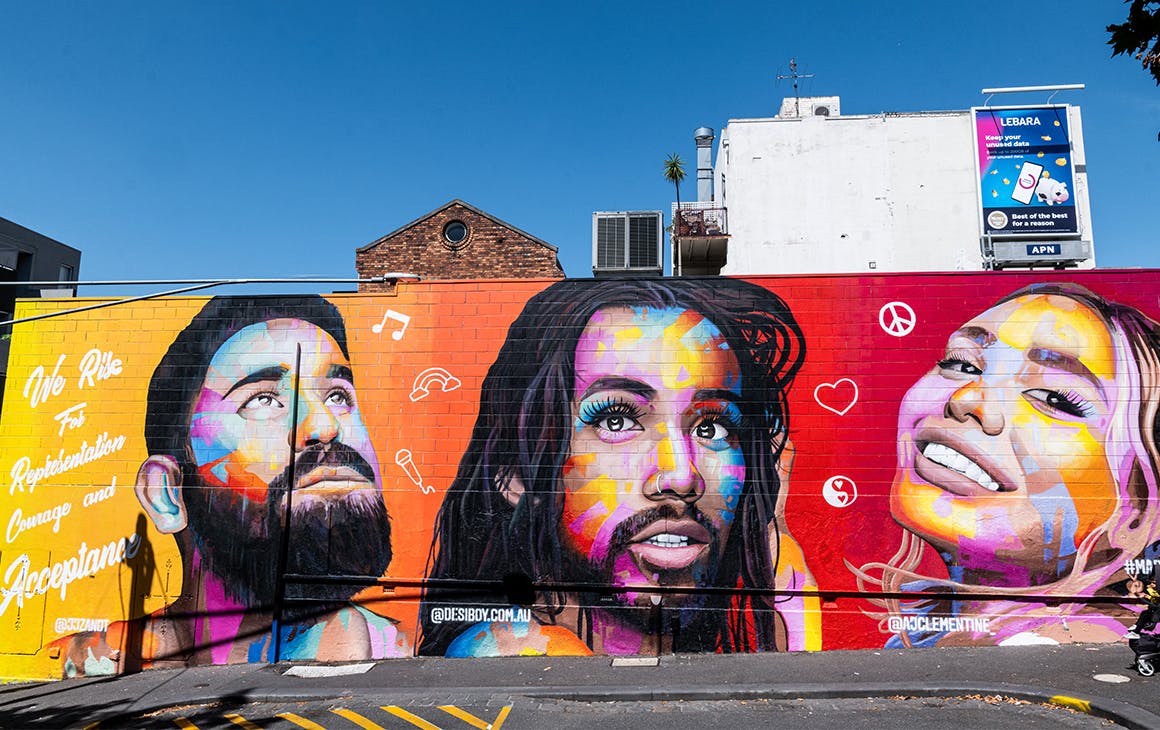 A colourful mural painted on a brick wall in Melbourne, a bright blue sky behind. The mural features the faces of AJ Clementine, Jeff van de Zandt and Roshan Nausad. 