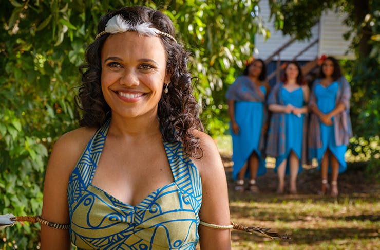 Image from Top End Wedding on Netflix. She smiles, while wearing a colourful dress. Behind her stands three women in blue dresses.
