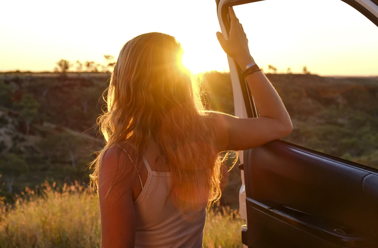 young woman facing sunset with hand on door of car