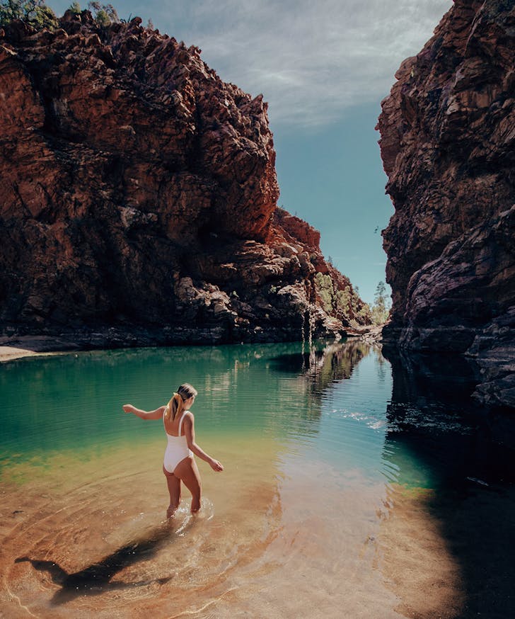 a woman walks into a strip of bright blue water, surrounded by a rocky gorge.