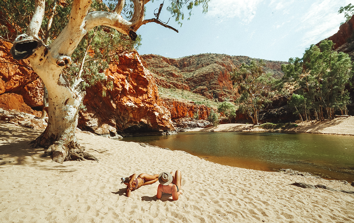a young man and woman recline on the sand next to natural pool at Ormiston Gorge.