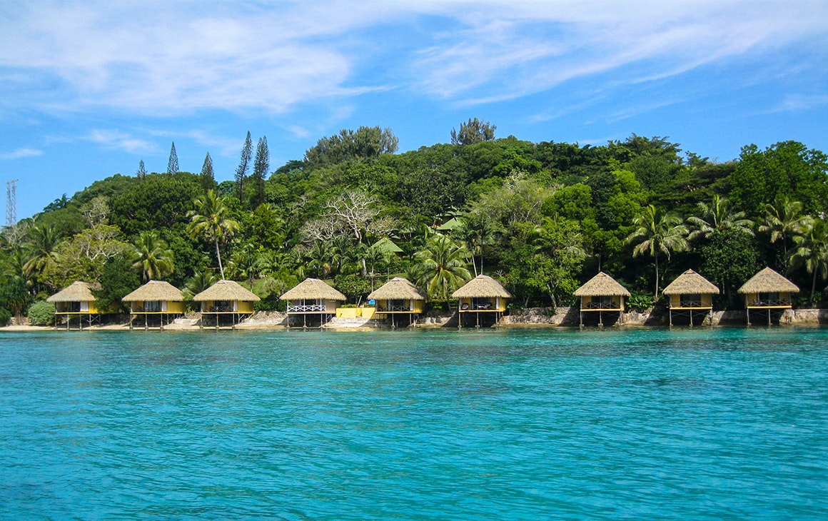 a line of wooden huts sit against a sparkling blue ocean