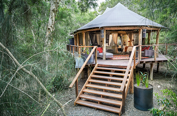 a gorgeous tent sits on a wooden deck in the middle of the bush.