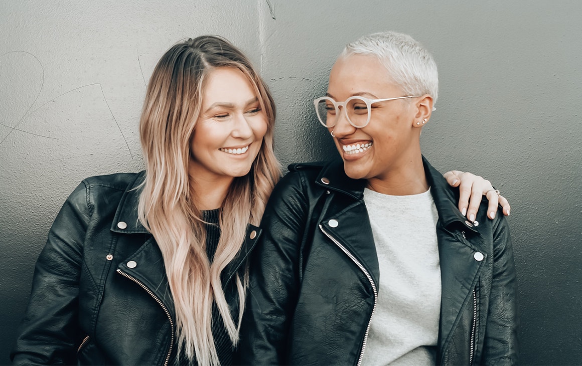 two women dressed in leather jackets smile and embrace.