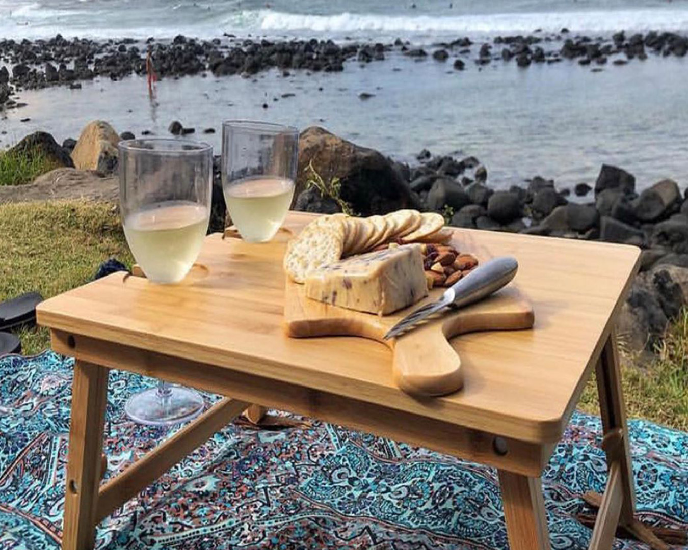 a small wooden picnic table with cheese, crackers and wine on it, sits next to the ocean.