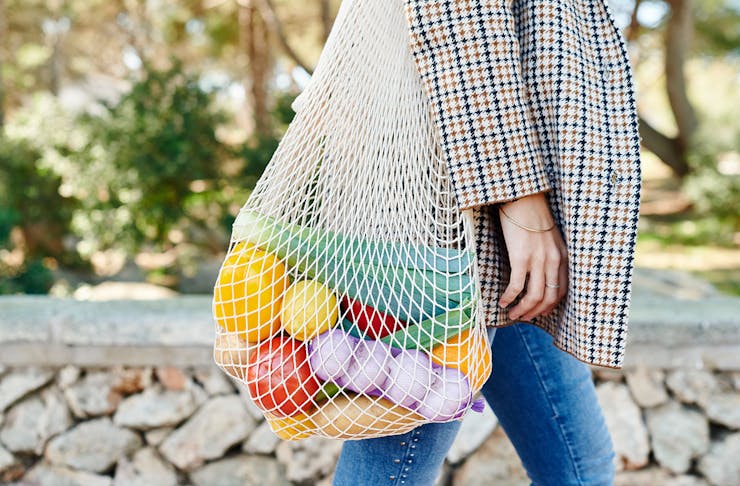 a woman has a reusable bag slung over her shoulder, filled with colourful fruit and veg. 