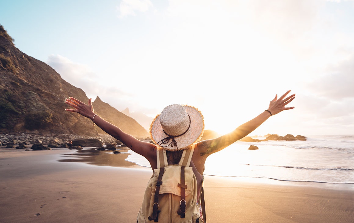 a woman stands on a beach at sunset wearing a hat and backpack. She's got her arms stretched out either side.