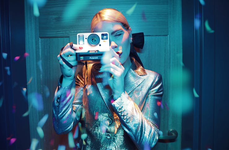 a woman, bathed in a blue light and surrounded by confetti, takes a photo on an old polaroid camera.