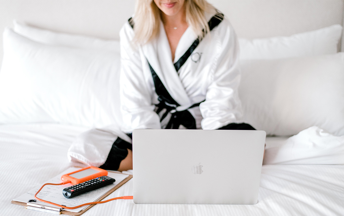 a woman, dressed in a white robe, sits on a plush, white bed while working on a laptop in front of her.