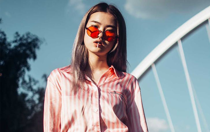 a young woman wearing orange tinted sunglasses, has a lollipop in her down. Behind her is blue sky and trees