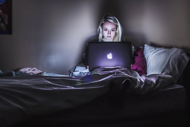 girl sitting on her bed in the dark, her face lit by light from open laptop