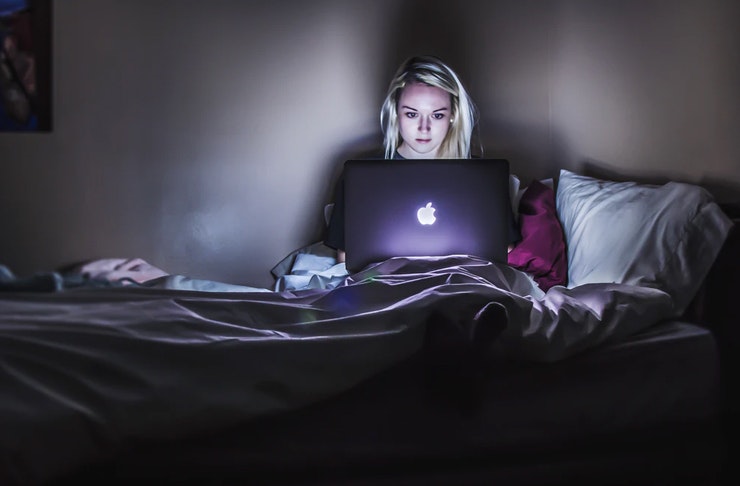 girl sitting on her bed in the dark, her face lit by light from open laptop