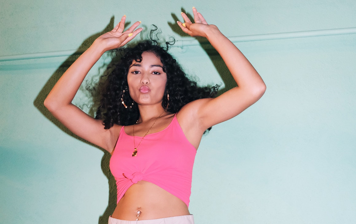 a woman in a pink crop top pouts and raises her arms in the air.