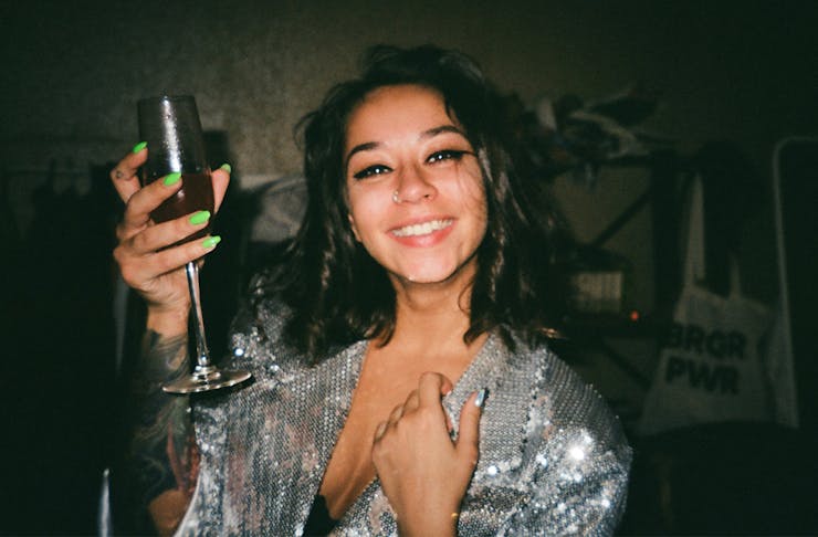 a woman in sparkly top raises a glass of bubble in the air, while smiling.