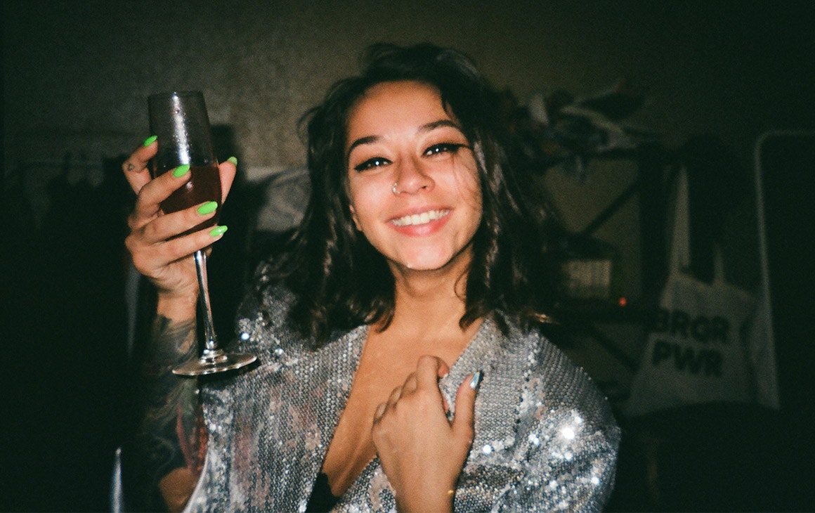 a woman in sparkly top raises a glass of bubble in the air, while smiling.