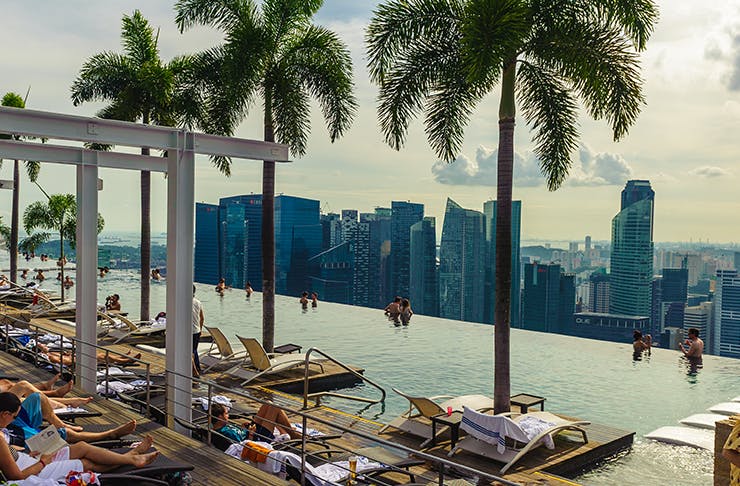 Rooftop infinity pool overlooking Singapore with people lounging by the pool under palm trees. 