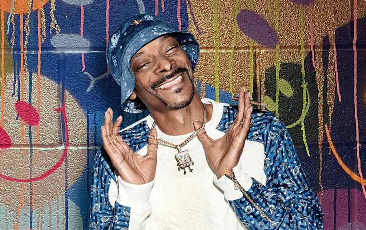 Snoop Dogg smiles, holding up his diamond necklace. He stands in front of a graffiti wall.