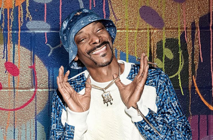 Snoop Dogg smiles, holding up his diamond necklace. He stands in front of a graffiti wall.