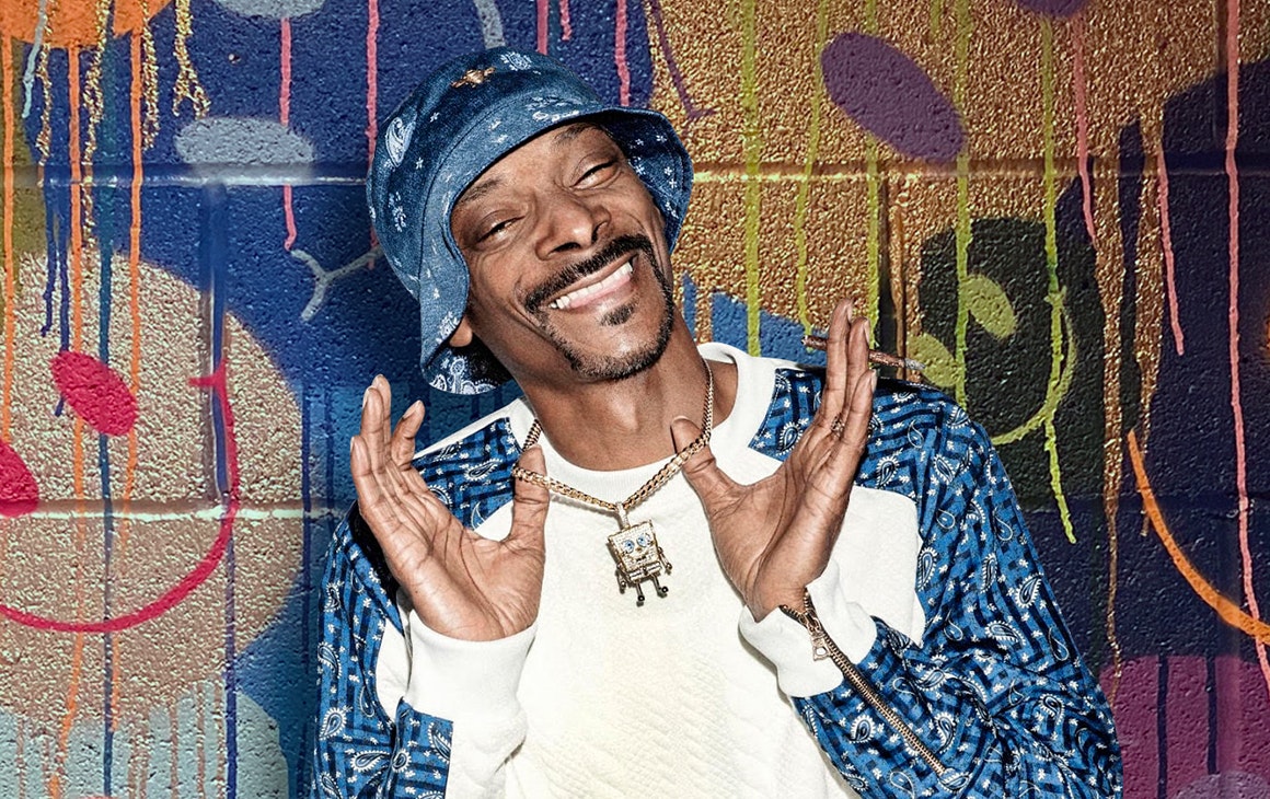 Snoop Dogg smiles, holding up his diamond necklace. He stands in front of a graffiti wall.