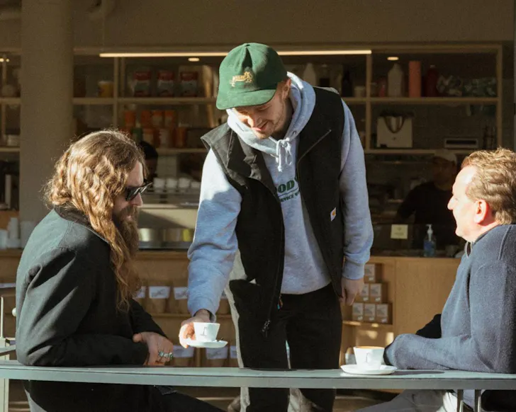Two people sit a cafe table in the sun, a third person places a coffee cup on the table