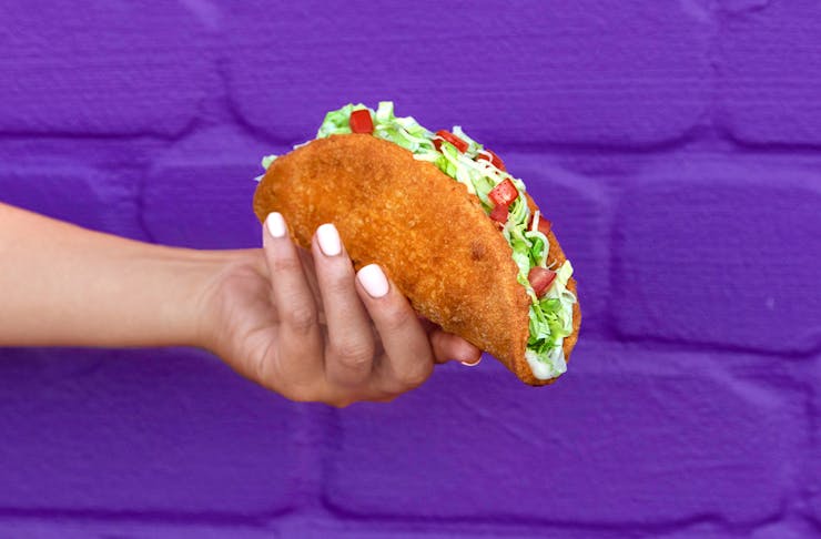A hand holding the naked chicken taco in front of a purple wall 