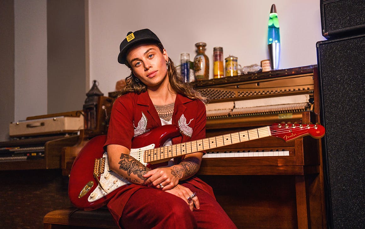 Tash Sultana sits at a piano, a guitar in their hands.