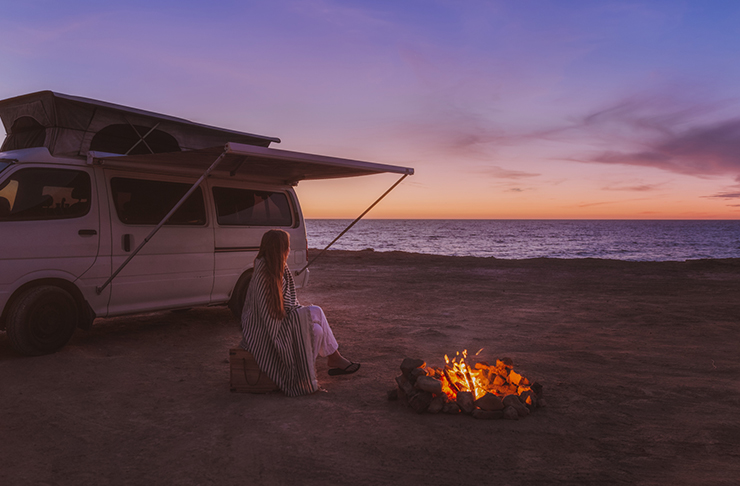 girl sitting on beach with van and fire at sunset