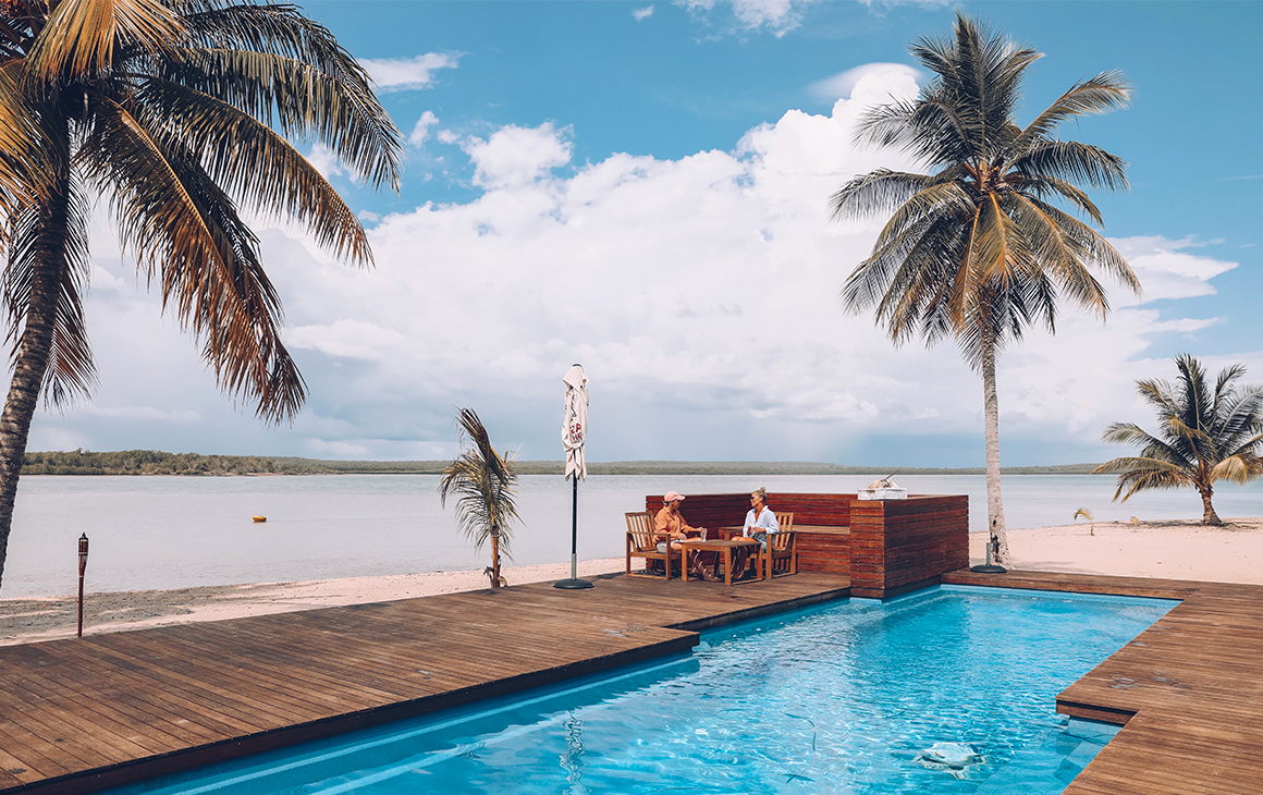 a couple sit beside a pool, fringed by palm trees at the Tiwi Island Retreat