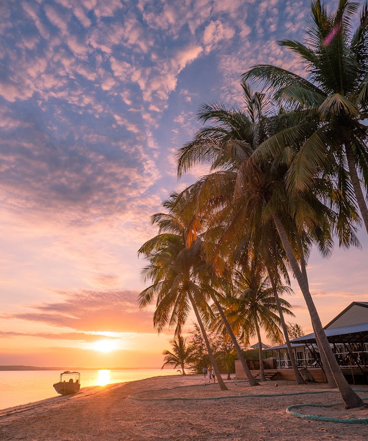 a dreamy sunset on Bathurst Island. There's a pink and purple sky, a boat in the distance and palm trees.