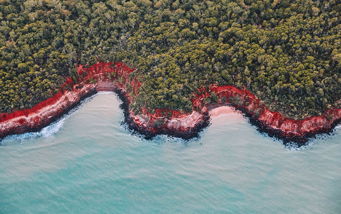 Stunning blue water meets a red sand beach, fringed by a lush bush.