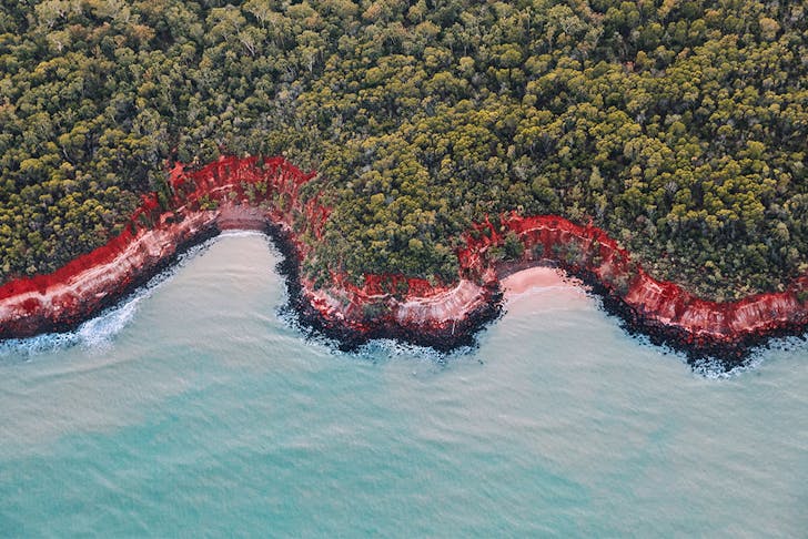 Stunning blue water meets a red sand beach, fringed by a lush bush.