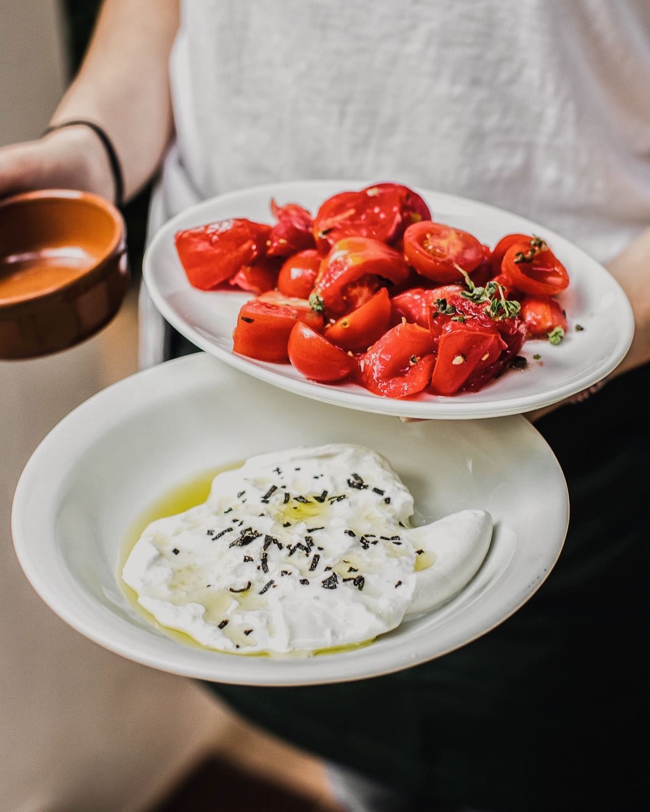 A person holds a bowl of cut cherry tomatoes and a burrata on a plate