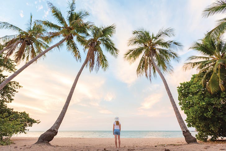 a woman walks on the beach, surrounded by towering palm trees