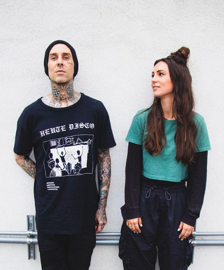 Travis Barker and Amy Shark lean against a white wall.
