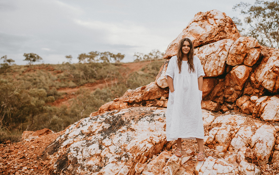 Glenda of Cungelela Art, poses on a red rock in the desert in a white dress and colourful TWOOBS.