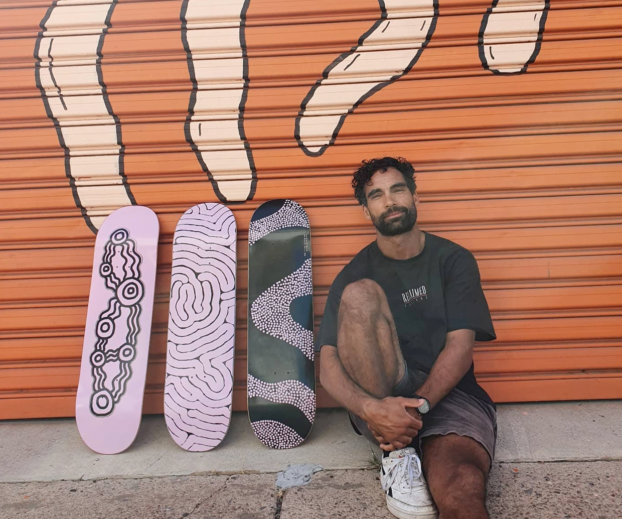 man leaning against garage door with three skateboards and his artwork on them